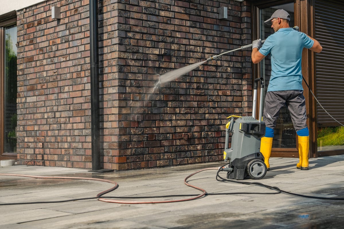 Man in boots and gloves operating a pressure washer cleaning a brick wall of a contemporary house