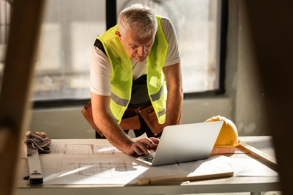 Construction worker in a bright vest managing tasks with plans and a laptop at a construction site